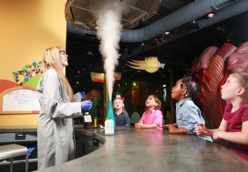 A scientist demonstrates a smoke effect to captivated children in a colorful educational setting.
