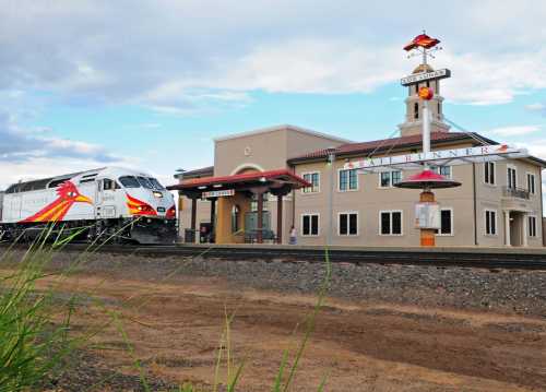 A train approaches a modern station with a red and yellow design, surrounded by grass and a cloudy sky.