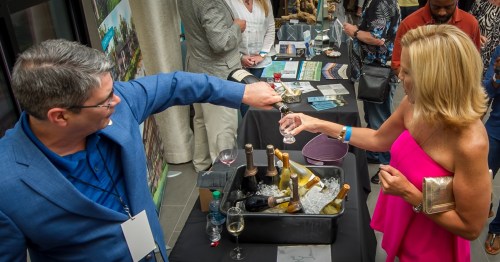 A man and woman toast with glasses at a social event, surrounded by tables with drinks and materials.