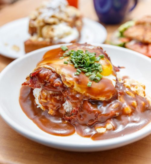 A bowl of rice topped with fried chicken, gravy, a fried egg, and green onions, served on a wooden table.