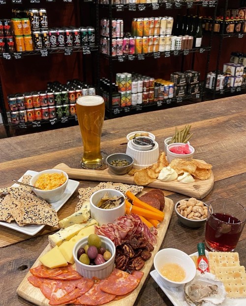A wooden table with a variety of charcuterie, cheeses, snacks, and drinks, set against a backdrop of shelves filled with cans.
