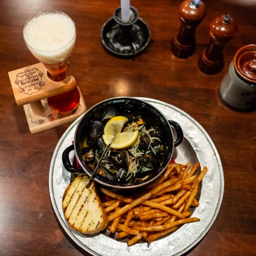A plate with mussels in broth, lemon, fries, and grilled bread, alongside a glass of beer on a wooden table.