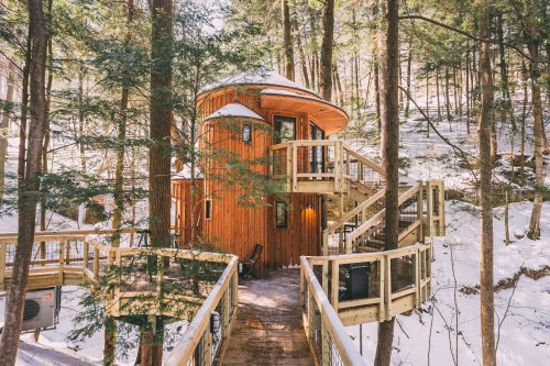 A cozy wooden treehouse surrounded by snow-covered trees, featuring a spiral staircase and a warm, inviting entrance.