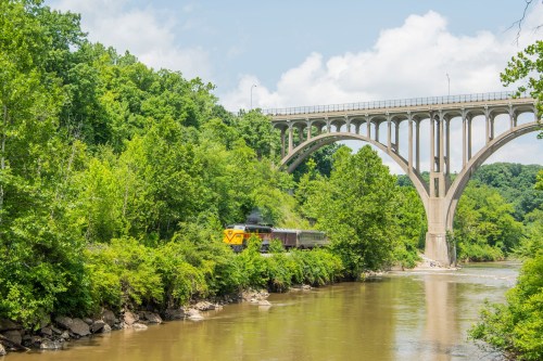 A train travels along a riverbank beneath a large, arched bridge surrounded by lush greenery.