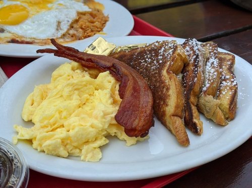 A plate of scrambled eggs, crispy bacon, and French toast dusted with powdered sugar, served with butter.