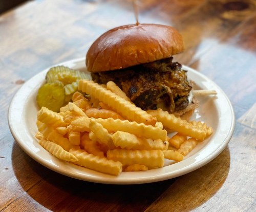 A plate with a pulled pork sandwich on a bun, served with crinkle-cut fries and pickles, on a wooden table.