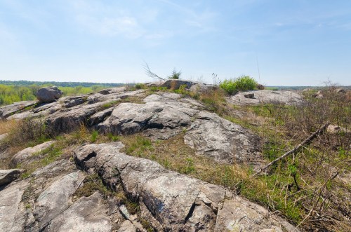 A rocky landscape with sparse vegetation under a clear blue sky, overlooking a distant green horizon.