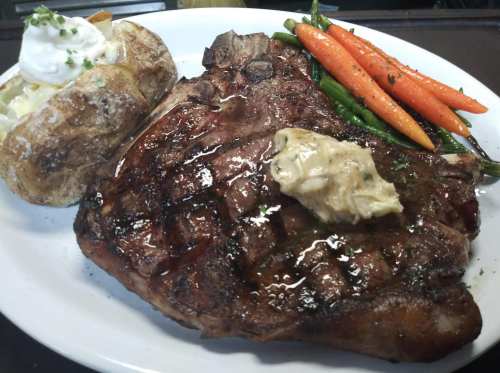 A large grilled steak topped with butter, served with a baked potato and colorful steamed carrots and green beans.