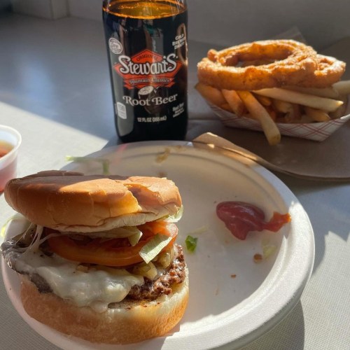 A plate with a burger topped with lettuce and tomato, fries, onion rings, and a bottle of root beer.