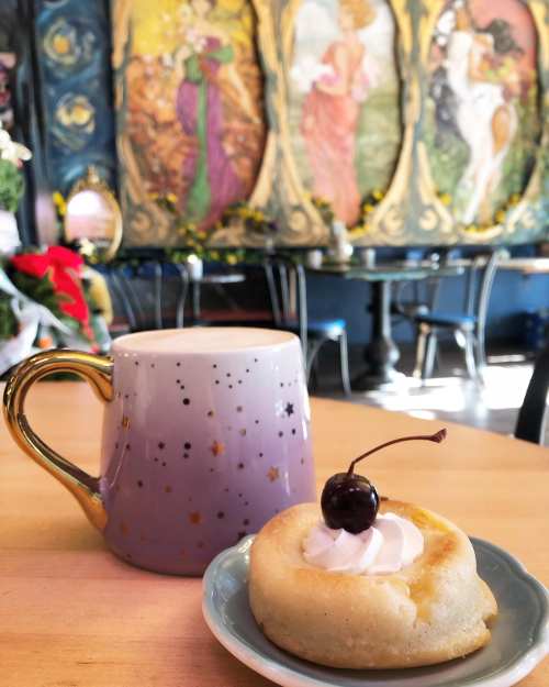 A cozy café scene featuring a purple starry mug and a dessert topped with whipped cream and a cherry, with colorful wall art.