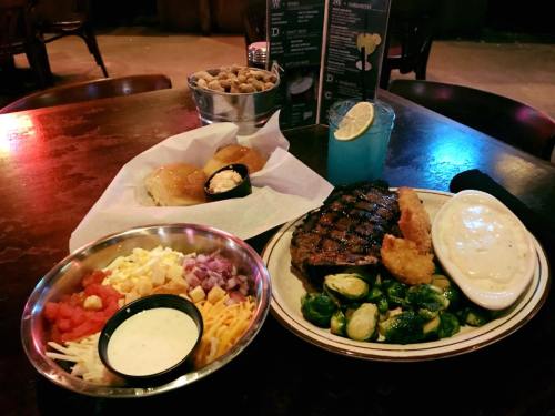 A table with a steak, shrimp, Brussels sprouts, salad, rolls, and a blue drink in a casual dining setting.