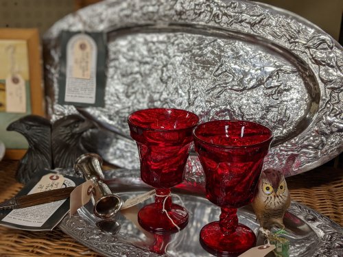 A collection of vintage items including red glass goblets, a silver platter, and decorative figurines on a woven surface.