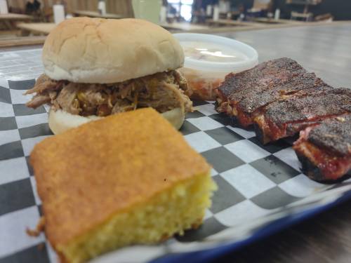 A plate featuring a pulled pork sandwich, cornbread, and a side of ribs with coleslaw, all on a checkered paper.