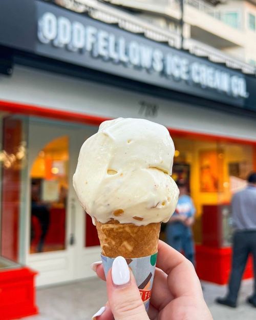 A hand holding a cone of creamy ice cream in front of Oddfellows Ice Cream Co. storefront.