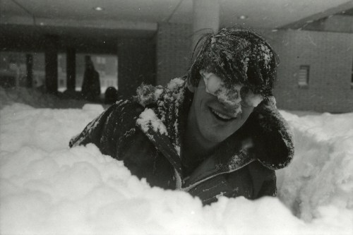 A person smiles while partially buried in deep snow, with snow covering their eyes and a snowy landscape around them.