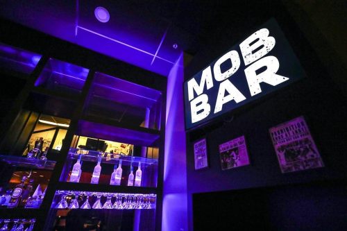 A dimly lit bar interior featuring a neon sign that reads "MOB BAR" and shelves with bottles and glasses.