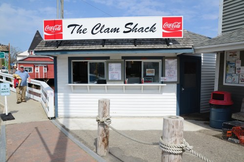 A small seafood shack with a Coca-Cola sign, featuring a takeout window and a rustic coastal setting.
