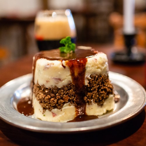 A layered dessert with mashed potatoes, chocolate sauce, and a crunchy topping, served on a metal plate.