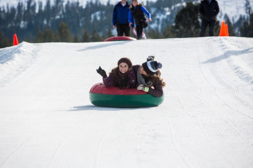 Two girls smile and wave while tubing down a snowy slope, with people in the background enjoying the winter fun.