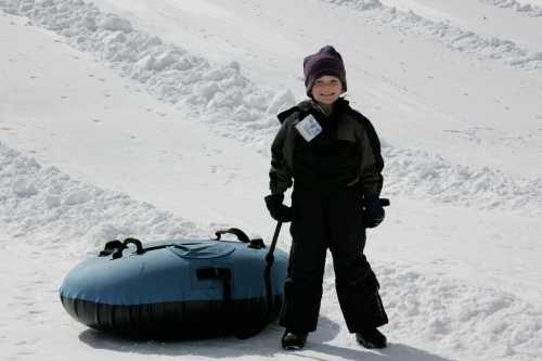 A smiling child in winter clothing stands next to a blue snow tube on a snowy slope.