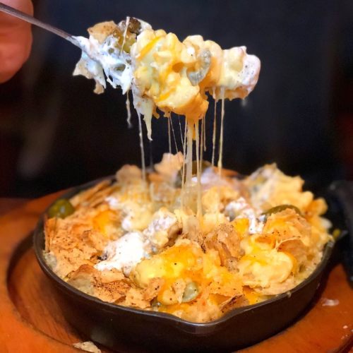 A hand holds a spoonful of cheesy, gooey nachos topped with sour cream and jalapeños, served in a skillet.