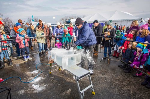 A crowd watches as a person cuts ice blocks with a chainsaw at an outdoor winter event.