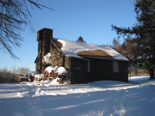 A rustic cabin covered in snow, with a chimney and surrounding trees under a clear blue sky.