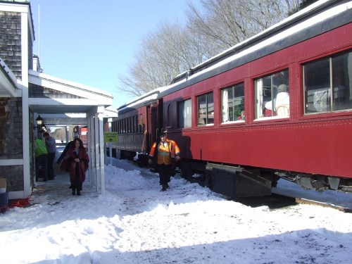 A red train parked near a snowy platform, with a person in an orange vest and a woman walking nearby.
