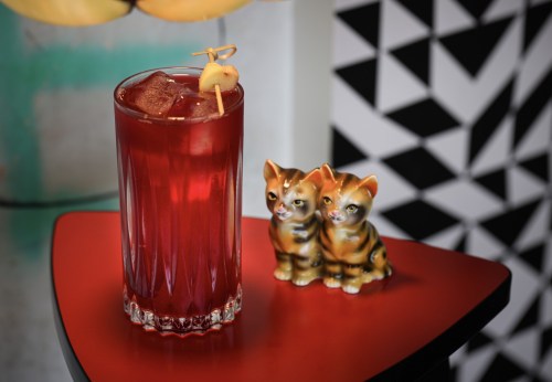 A vibrant red cocktail with ice sits on a red table next to two small ceramic cat figurines.