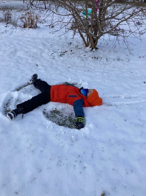 A child in an orange jacket and hat lies in the snow, making snow angels in a winter landscape.