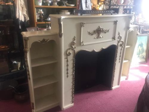 A vintage white fireplace mantel with ornate gold accents and side shelves, set in a cozy antique shop.