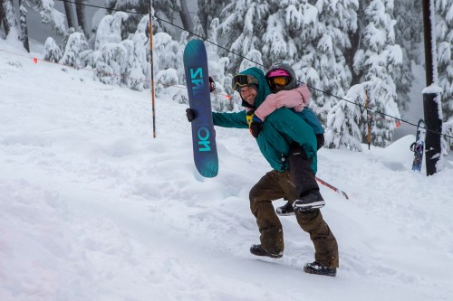 A person carries a child on their back while holding a snowboard, surrounded by snowy trees.