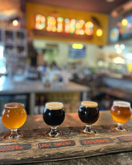 A wooden flight board with four different craft beers in small glasses, set against a blurred bar background.