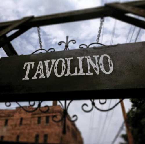 Sign reading "TAVOLINO" hanging from a decorative structure against a cloudy sky.