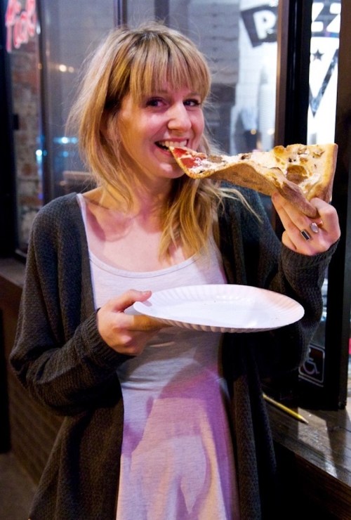 A smiling woman holds a large slice of pizza over a paper plate, ready to take a bite, outside a pizzeria at night.