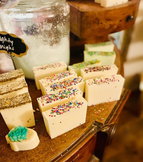 A display of colorful handmade soaps on a wooden shelf, featuring various shapes and decorative toppings.