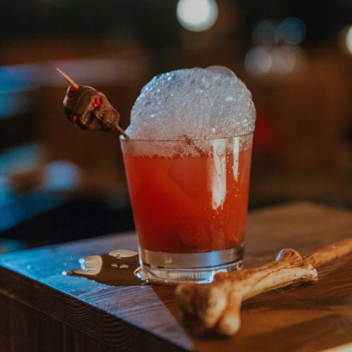 A vibrant red cocktail with foam, garnished with a skewer, sits on a wooden surface with a bone beside it.