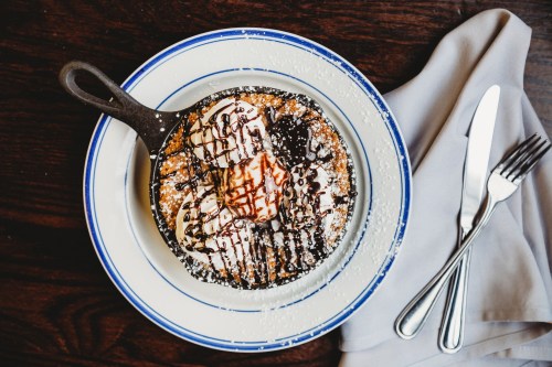 A skillet dessert topped with ice cream, chocolate drizzle, and powdered sugar, served on a plate with utensils.