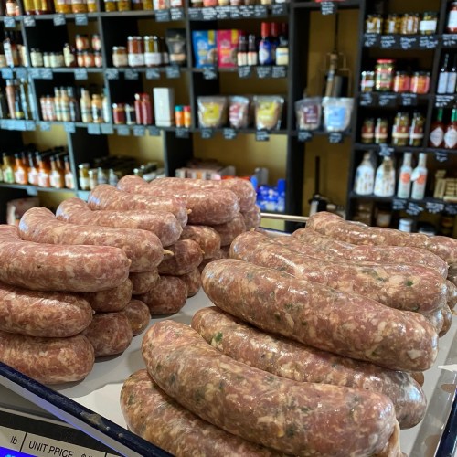 A display of fresh sausages stacked on a counter, with shelves of sauces and condiments in the background.