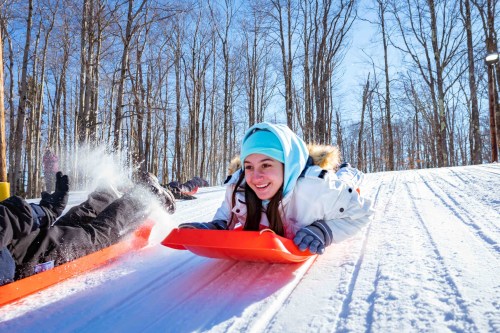 A girl in a winter jacket and blue hat sleds down a snowy hill, smiling as she enjoys the winter fun.