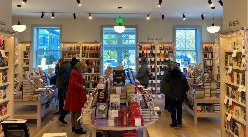 A cozy bookstore with shelves filled with books and customers browsing among the displays. Natural light fills the space.