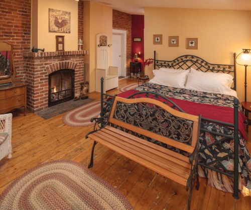 Cozy bedroom with a brick fireplace, wooden floors, a decorative bed, and a vintage bench. Warm, inviting atmosphere.