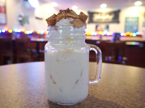 A glass mason jar filled with a creamy dessert topped with cereal, set on a table in a softly lit bar.