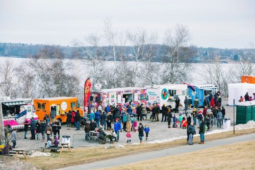 A crowd gathers at food trucks by a lake, with trees and a cloudy sky in the background.