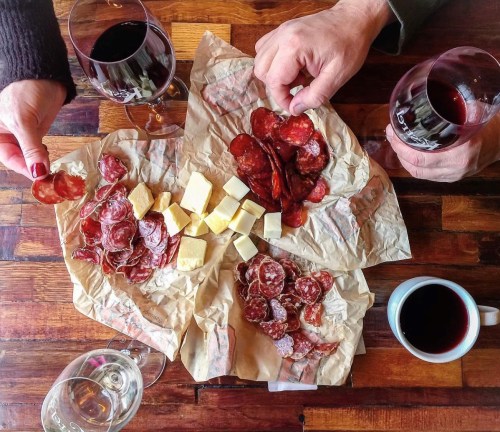 A wooden table with wine glasses, a coffee cup, and a platter of assorted meats and cheese on crumpled paper.
