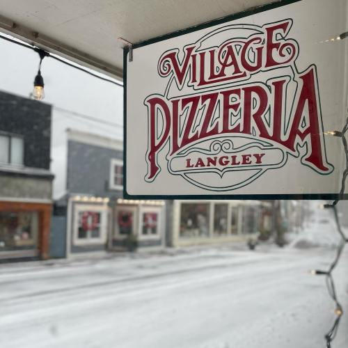 Sign for "Village Pizzeria" in Langley, with a snowy street scene visible in the background.