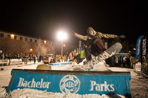 A snowboarder performs a trick on a rail during a night event, with a crowd and lights in the background.