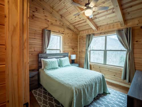 Cozy wooden bedroom with a queen bed, large windows, and a ceiling fan, featuring natural light and a serene atmosphere.