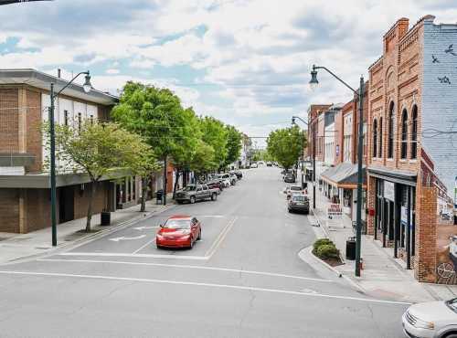 A quiet street scene with trees lining the road, parked cars, and buildings on either side under a cloudy sky.