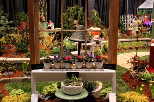 A garden display featuring colorful flowers, decorative plants, and a wooden frame with potted plants in front.